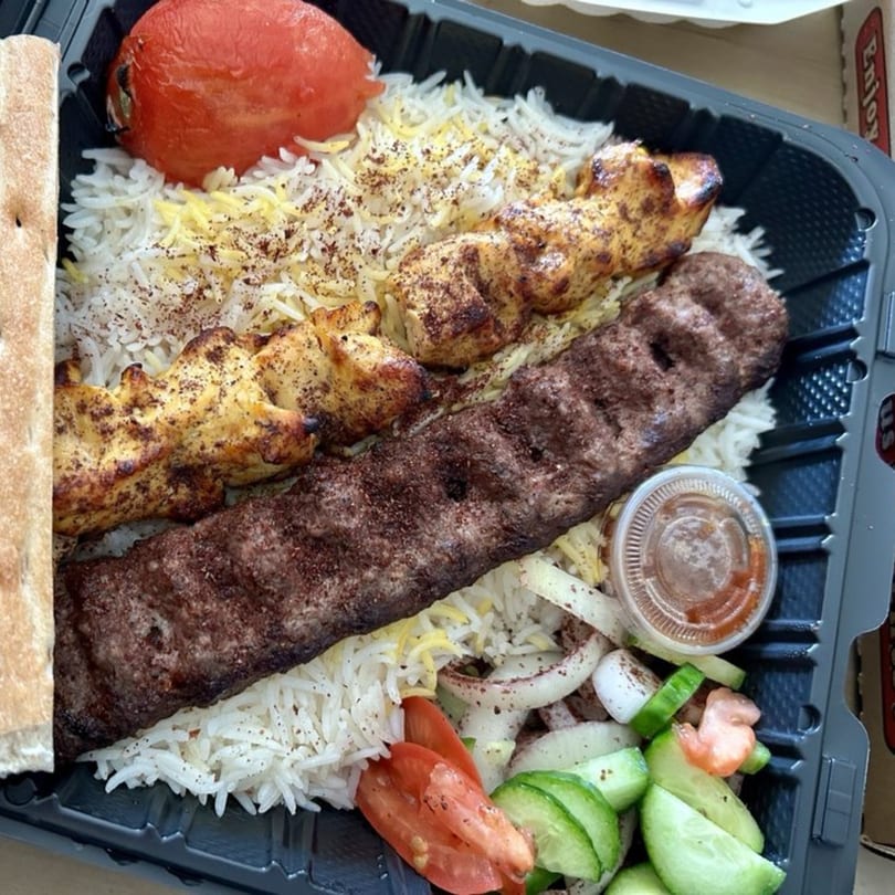 Halal chicken and beef kebabs with rice, grilled tomato, pita bread, and salad in a takeout container.