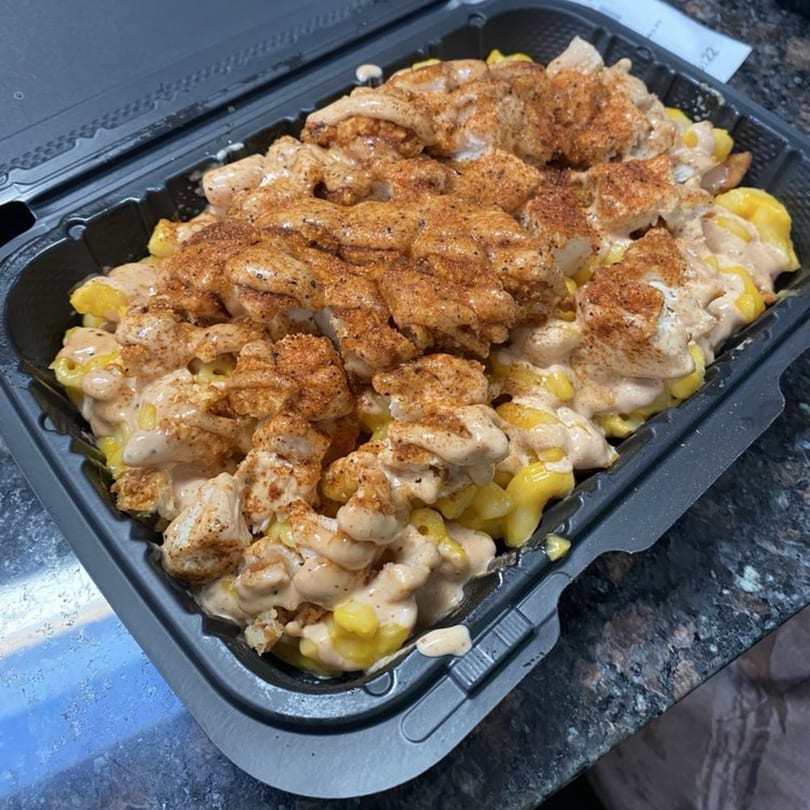Halal mac and cheese topped with seasoned chicken pieces and creamy sauce in a black takeout container.